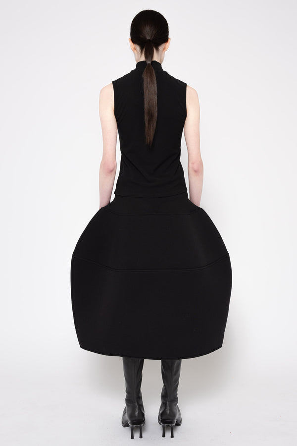 A model shown from the back wearing a black sleeveless dress with a high neck and a very large, voluminous skirt, paired with black knee-high boots against a plain white studio background.