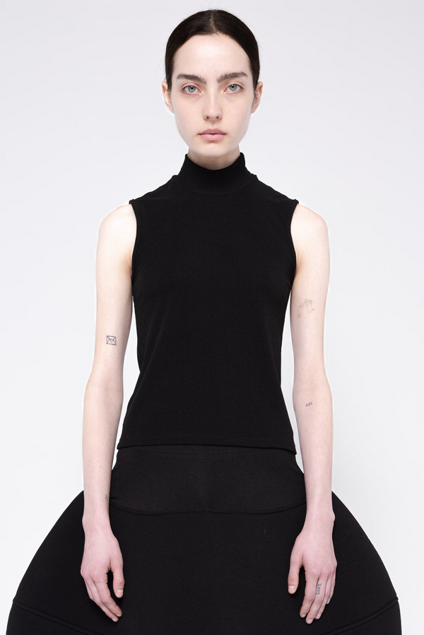 A model wearing a black sleeveless mock-neck top and a matching black skirt, photographed against a light gray studio background.