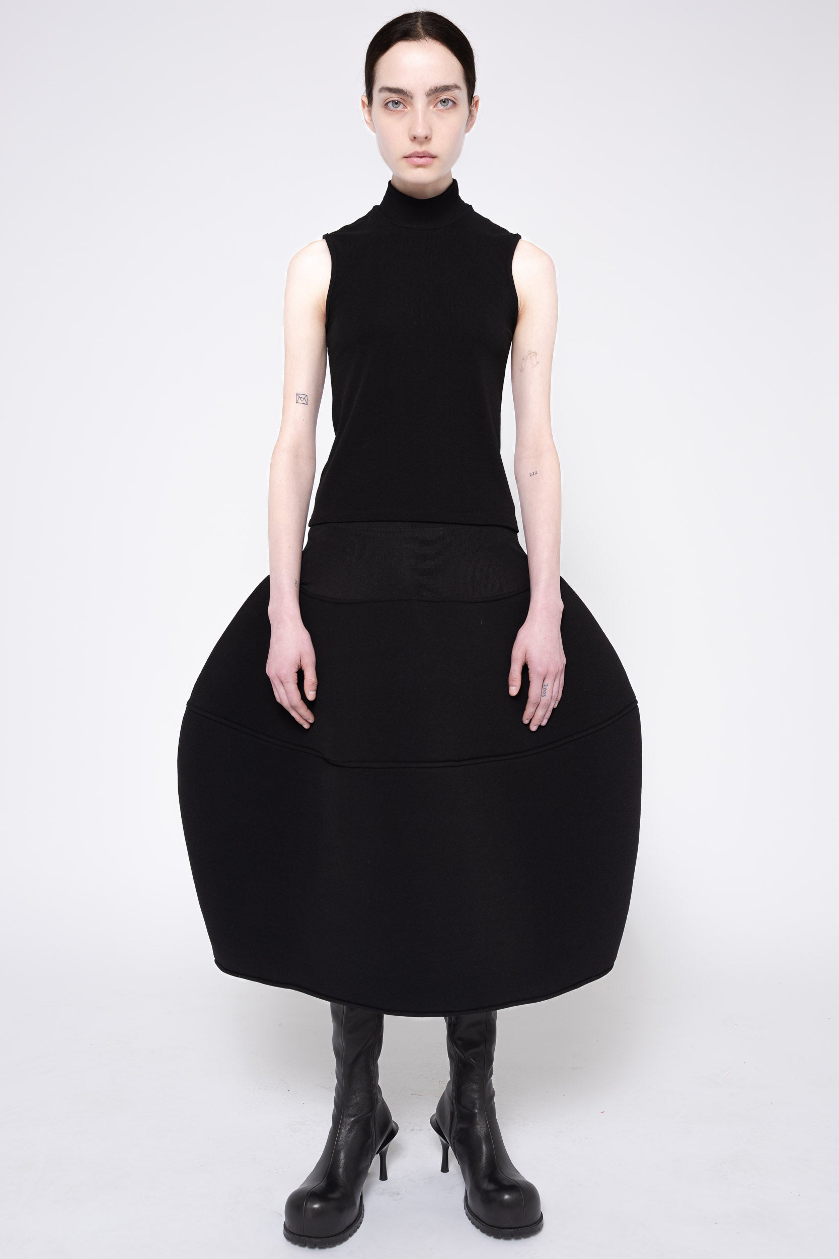 A model wearing a black sleeveless mock-neck dress with a very large, structured balloon skirt and black heeled boots, photographed against a light gray studio background.
