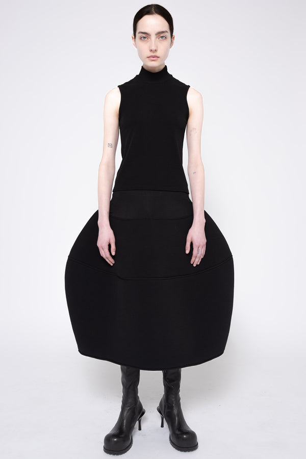A model wearing a black sleeveless mock-neck dress with a very large, structured balloon skirt and black heeled boots, photographed against a light gray studio background.