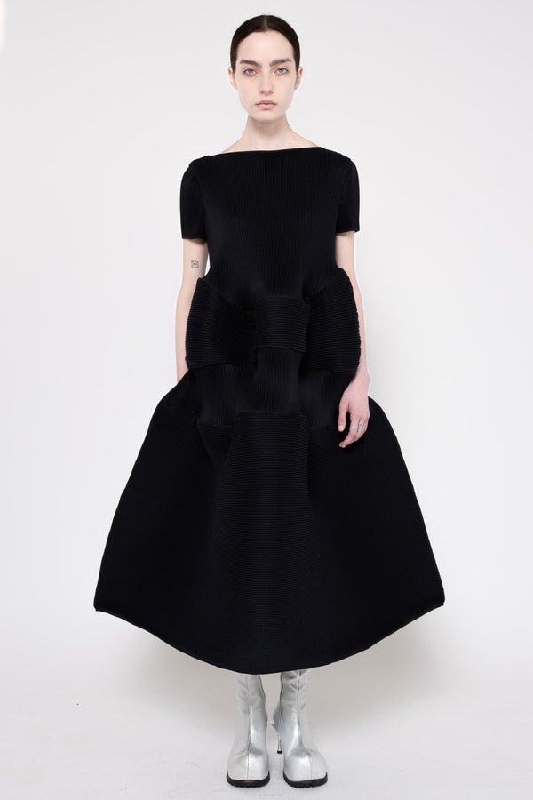 A model wearing a black short-sleeved midi dress with a structured, voluminous skirt and silver metallic boots, standing against a plain white studio background.