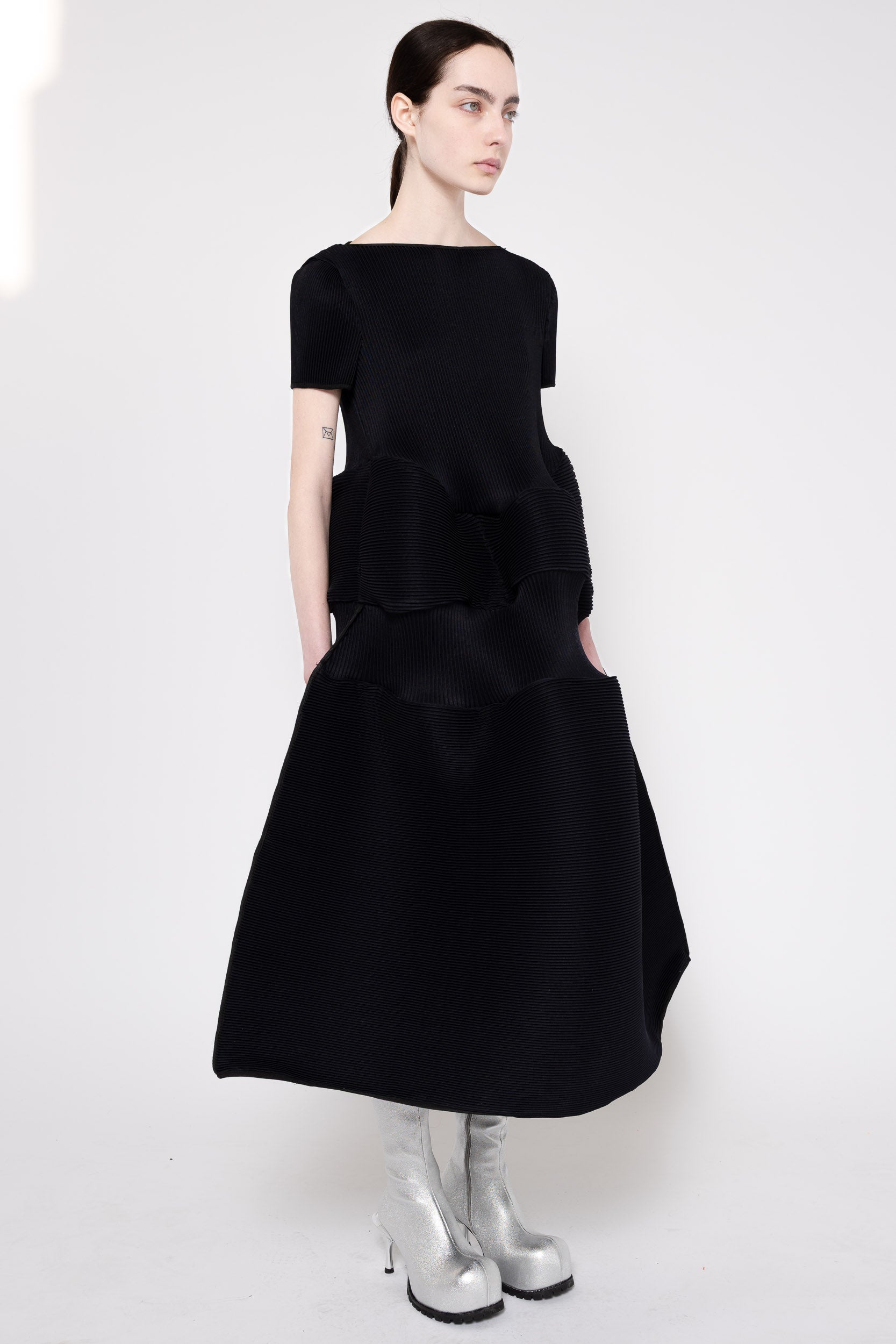 A model wearing a black short-sleeved midi dress with structured, tiered ruffle layers and silver metallic knee-high boots, photographed against a plain white studio background.