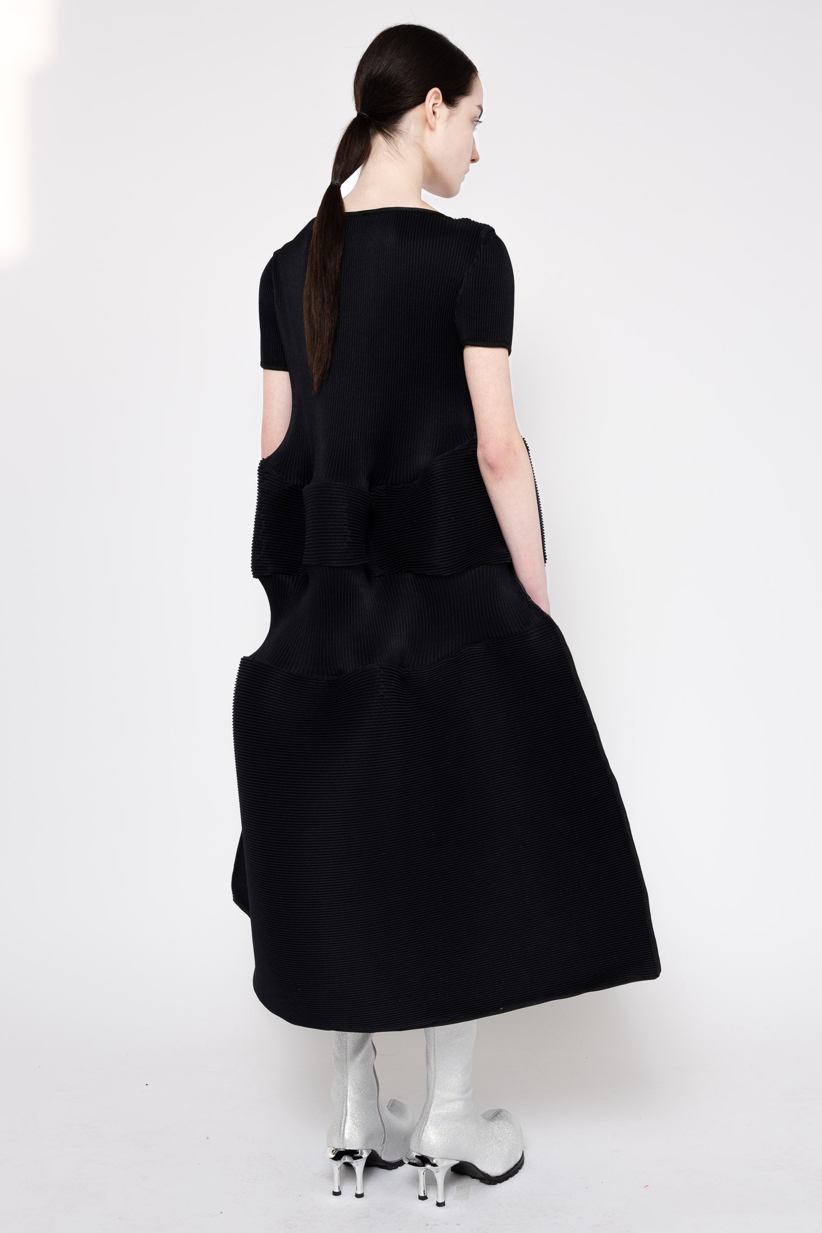 A model shown from the back wearing a black short-sleeved midi dress with a structured, tiered ruffled back design and pockets, paired with white high-heeled boots against a plain white studio background.
