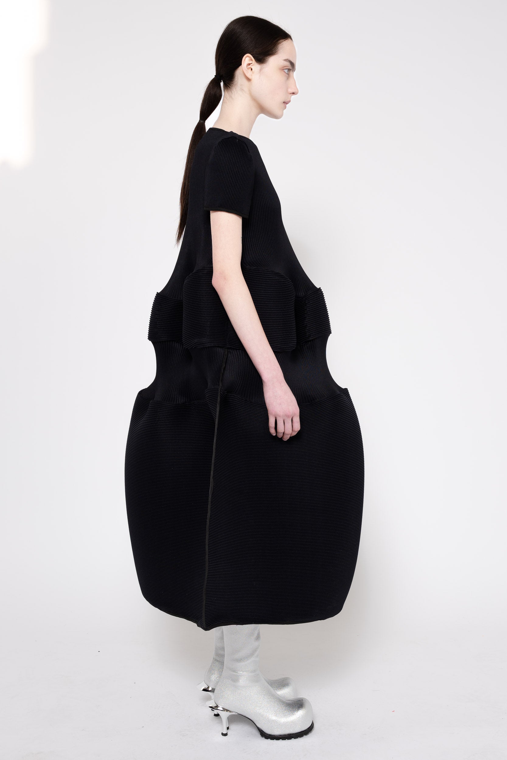 A model shown from the back wearing a black short-sleeve dress with a large structured, balloon-like skirt and silver high-heeled boots against a plain white studio background.