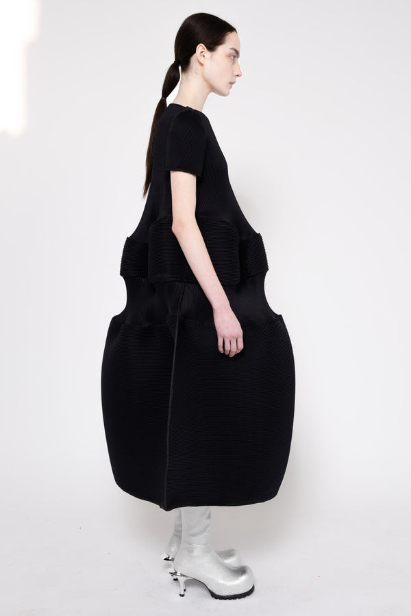 A model shown from the back wearing a black short-sleeve dress with a large structured, balloon-like skirt and silver high-heeled boots against a plain white studio background.