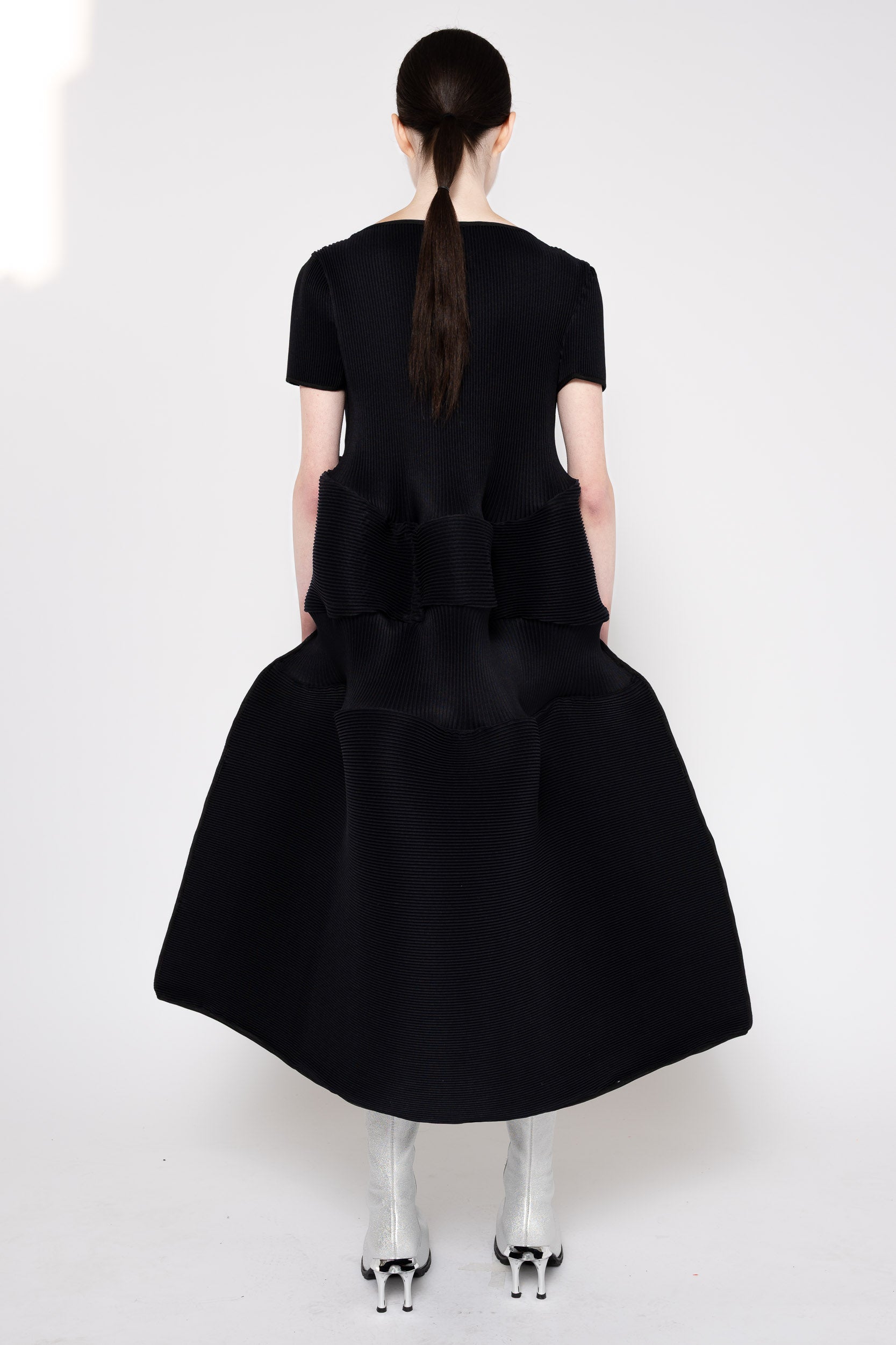 A model shown from the back wearing a black short-sleeve dress with a dramatic, oversized structured waist and voluminous skirt, paired with metallic silver high-heeled shoes against a plain white studio background.