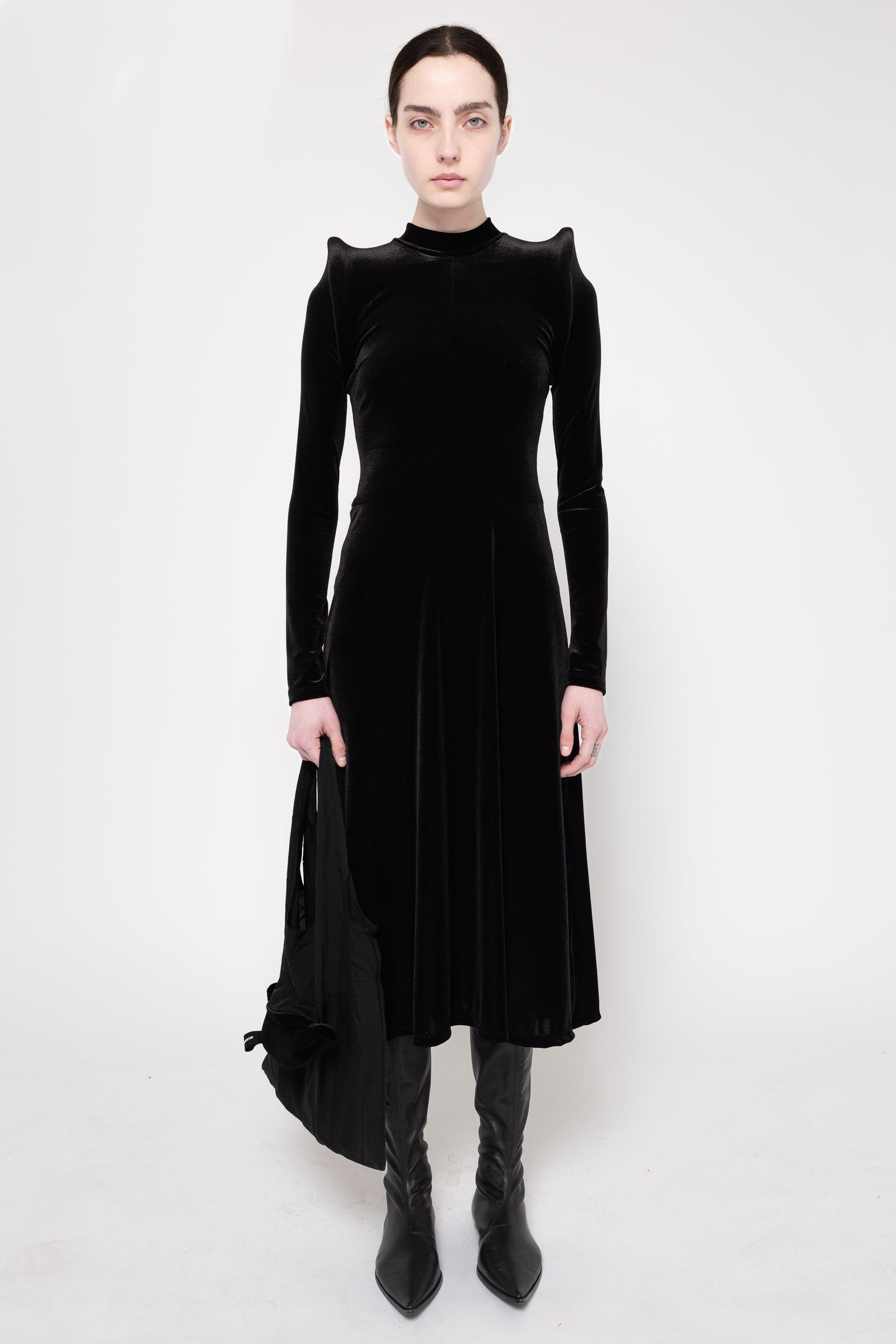 A model wearing a black velvet long-sleeve midi dress with dramatic shoulder ruffles, paired with black pointed-toe ankle boots, against a plain white studio background.