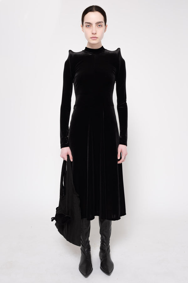 A model wearing a black velvet long-sleeve midi dress with dramatic shoulder ruffles, paired with black pointed-toe ankle boots, against a plain white studio background.