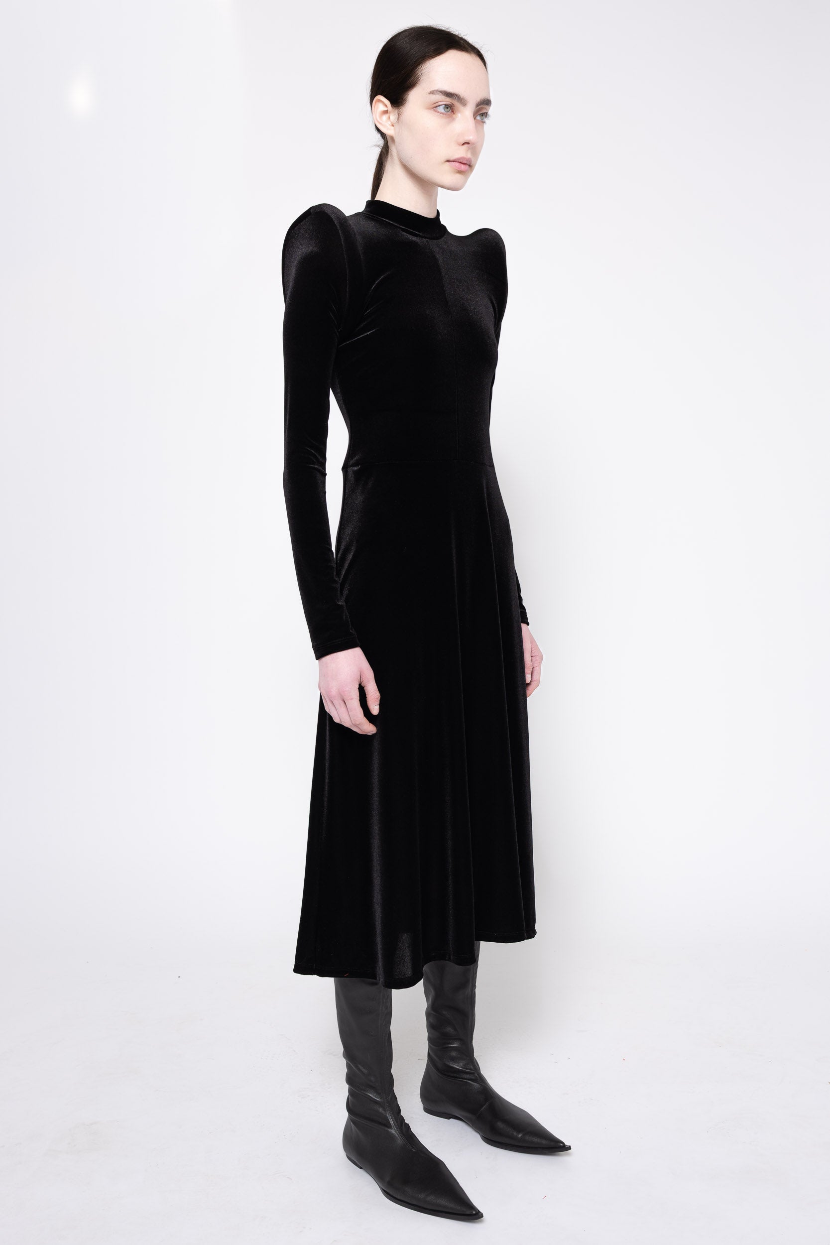 A model wearing a black velvet long-sleeve midi dress with structured shoulders, paired with black pointed-toe boots, photographed against a plain white studio background.