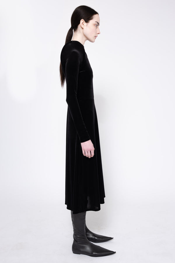 A model in profile wearing a long-sleeved black velvet midi dress, black pointed-toe boots, and black socks against a plain white studio background.