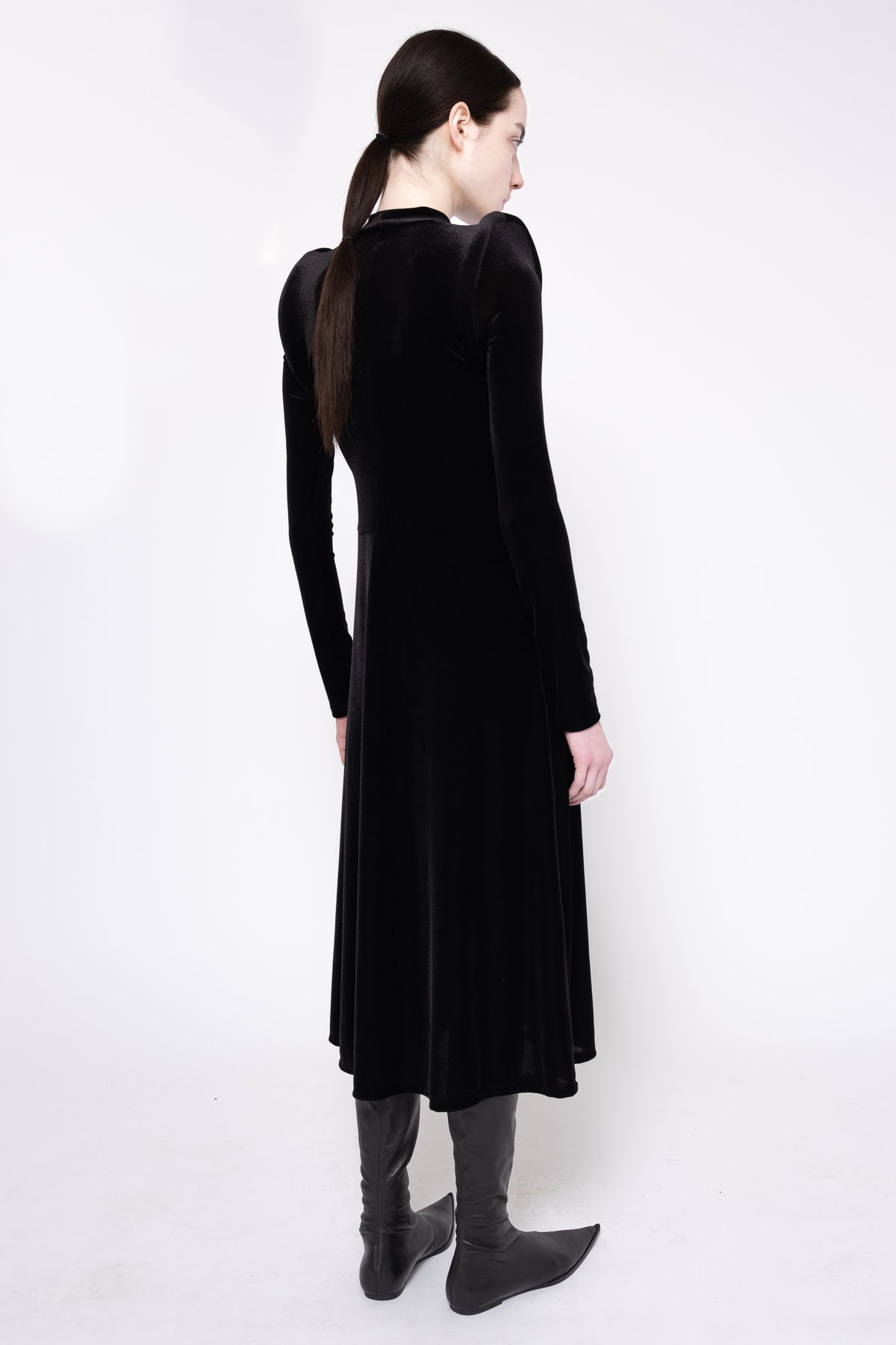 A woman wearing a long black velvet long-sleeve dress, shown from the back, paired with dark gray boots, against a plain white studio background.