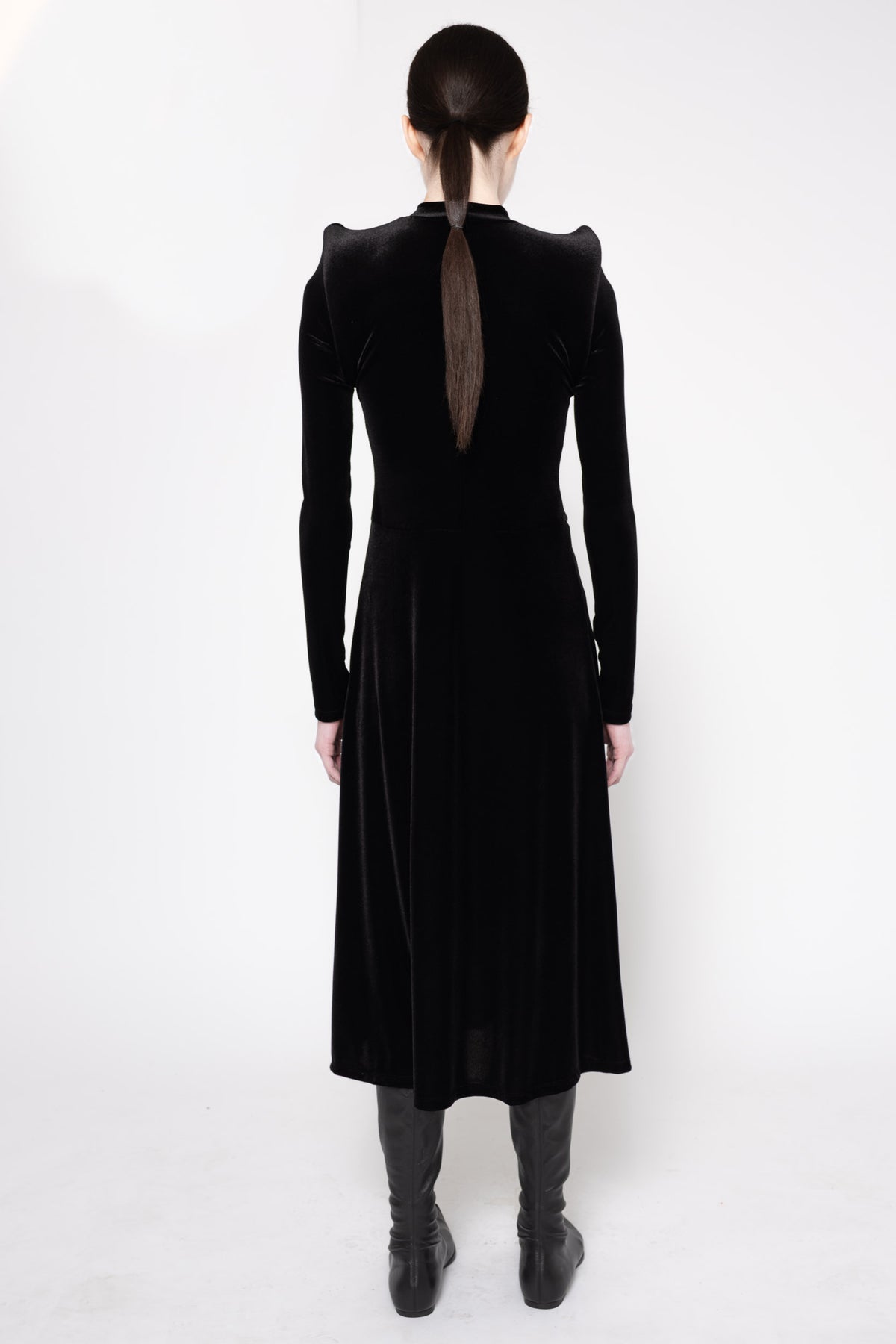 A model viewed from the back wearing a long black velvet dress with structured shoulders and a sheer back panel, paired with dark gray boots against a white studio background.