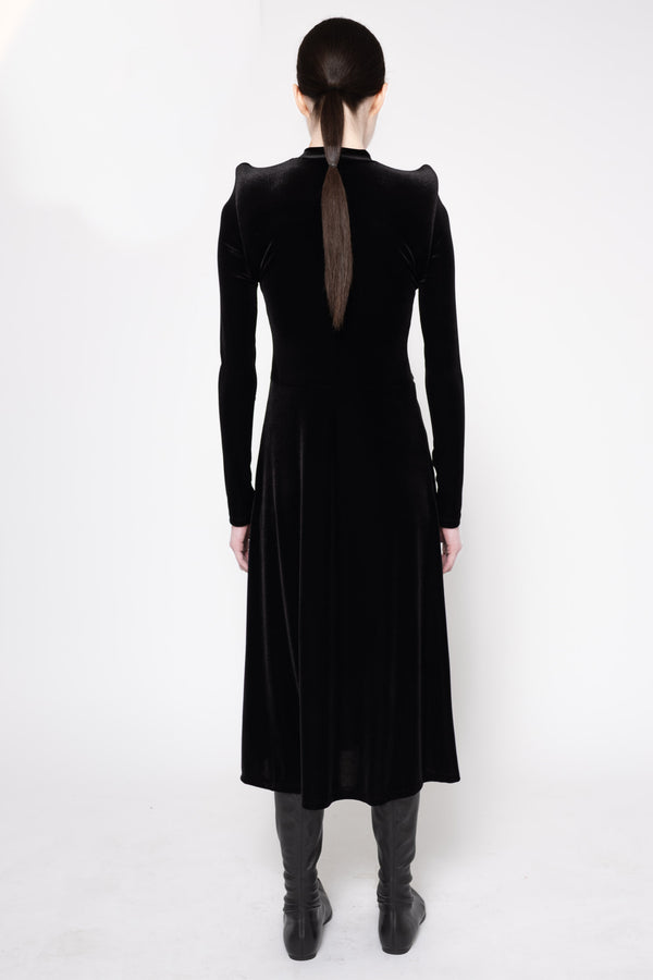A model viewed from the back wearing a long black velvet dress with structured shoulders and a sheer back panel, paired with dark gray boots against a white studio background.