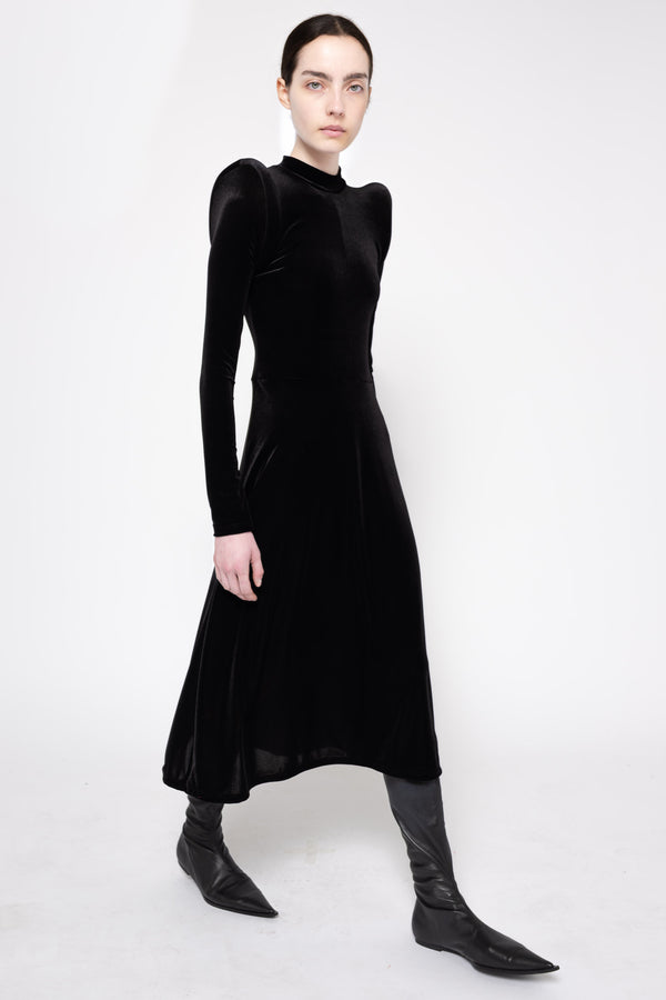 A model wearing a black velvet long-sleeve midi dress with structured shoulders, paired with black pointed-toe boots, against a plain white studio background.