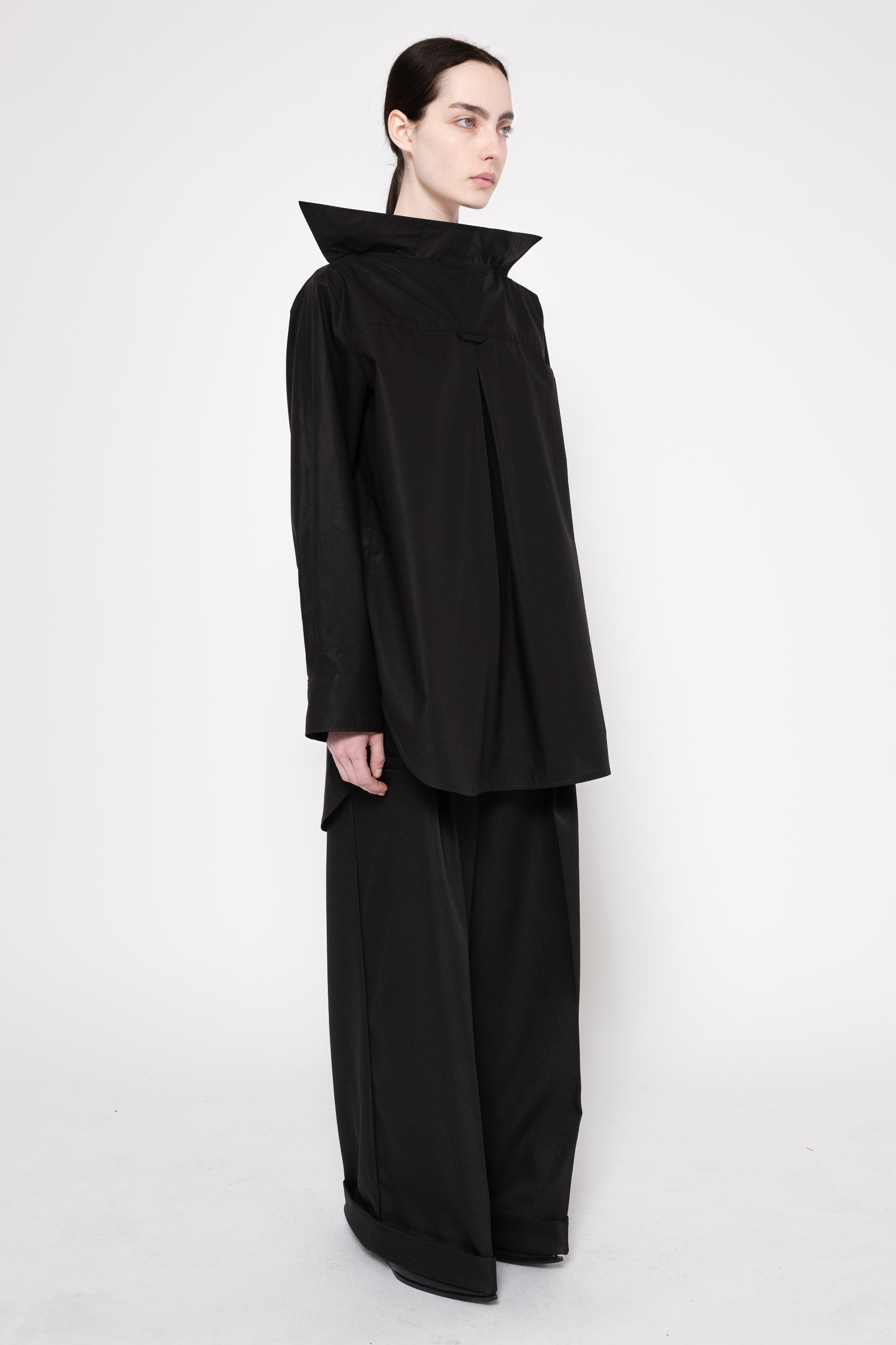 A model wearing a black asymmetrical long-sleeve top with a dramatic high collar and matching wide-leg trousers, photographed against a seamless white studio background.