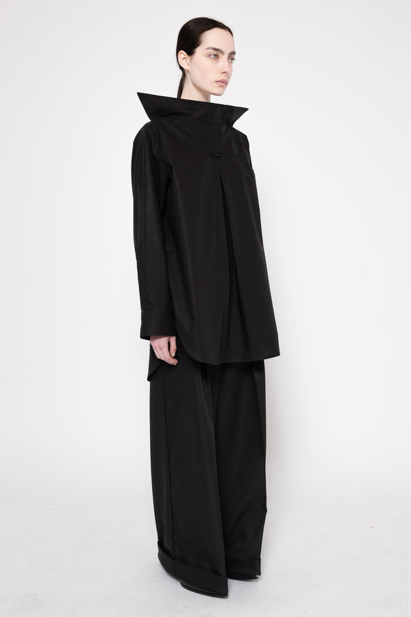 A model wearing a black asymmetrical long-sleeve top with a dramatic high collar and matching wide-leg trousers, photographed against a seamless white studio background.