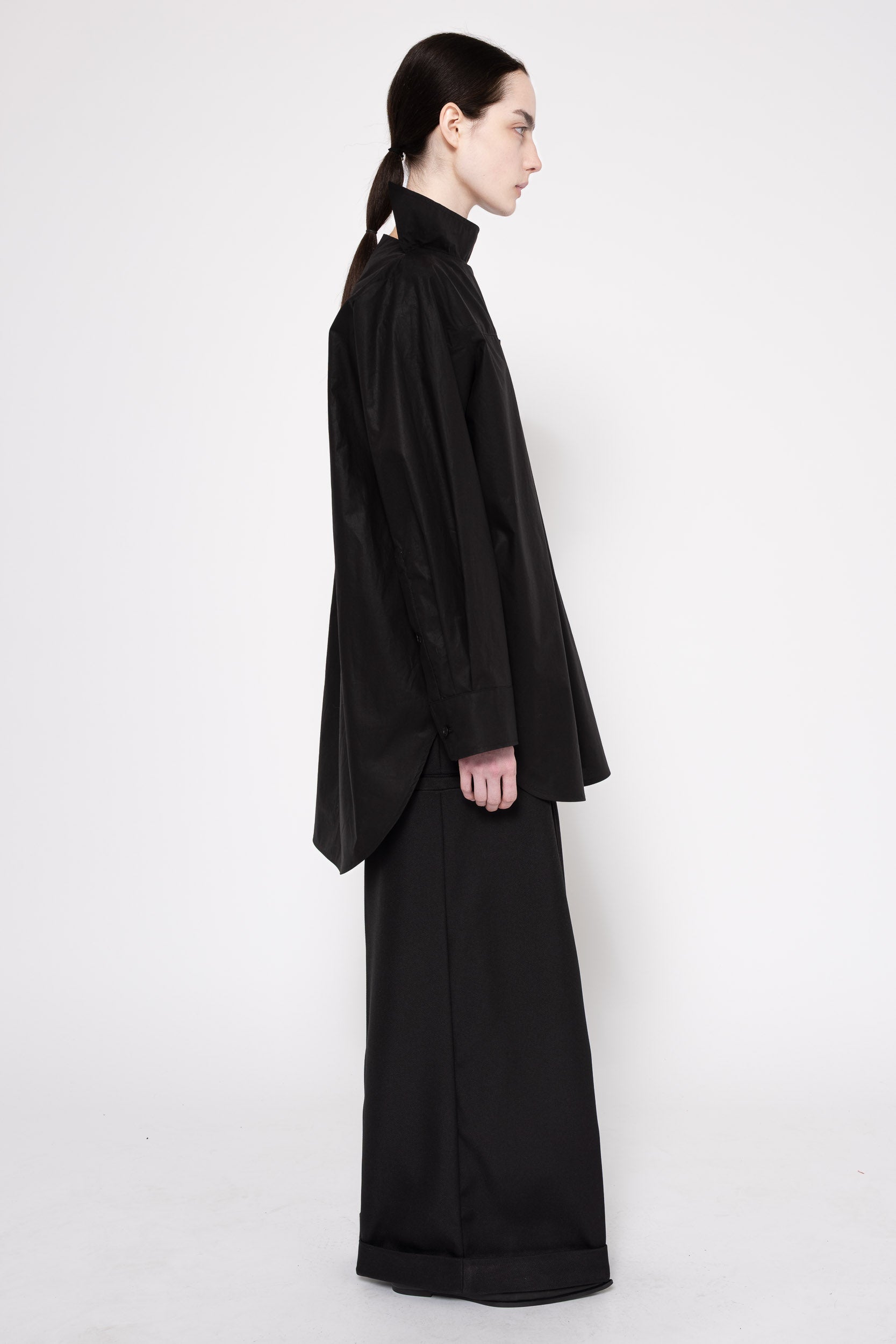A model in profile wearing a black high-neck, oversized long-sleeve top and matching wide-leg trousers against a plain light gray studio background.