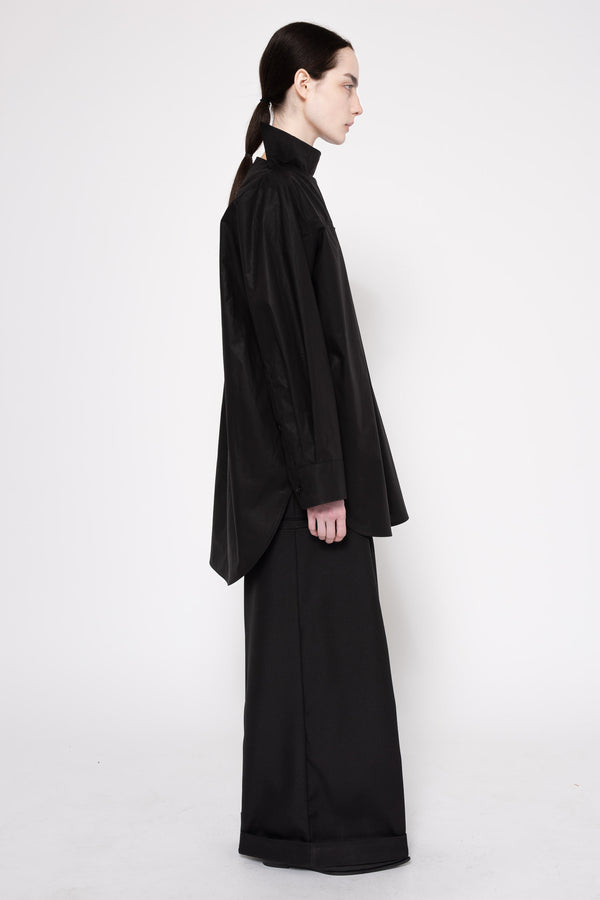 A model in profile wearing a black high-neck, oversized long-sleeve top and matching wide-leg trousers against a plain light gray studio background.