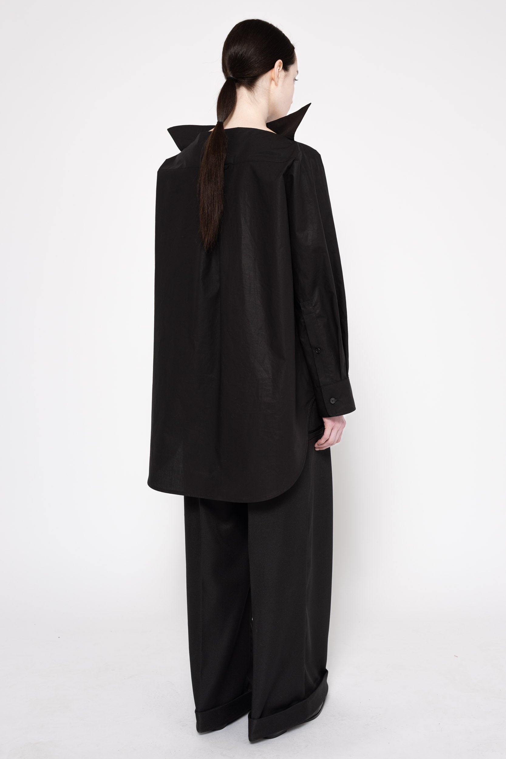 A model shown from the back wearing a black long-sleeve top with dramatic pointed collar details and matching wide-leg black trousers, set against a plain white studio background.