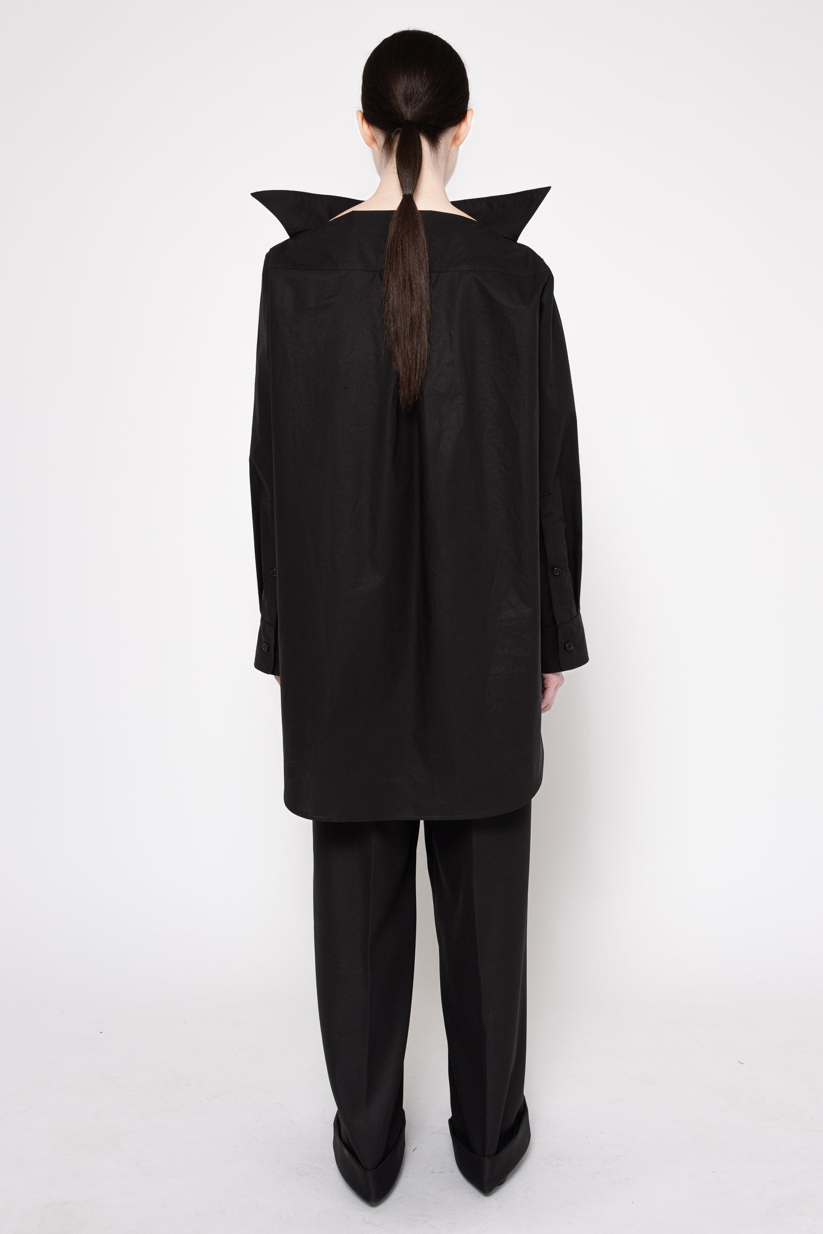 A model shown from the back wearing a black oversized long-sleeve top with dramatic pointed shoulder details and matching black wide-leg trousers, set against a plain white studio background.