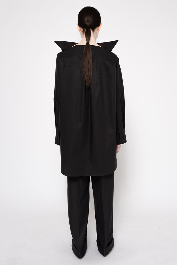 A model shown from the back wearing a black oversized long-sleeve top with dramatic pointed shoulder details and matching black wide-leg trousers, set against a plain white studio background.