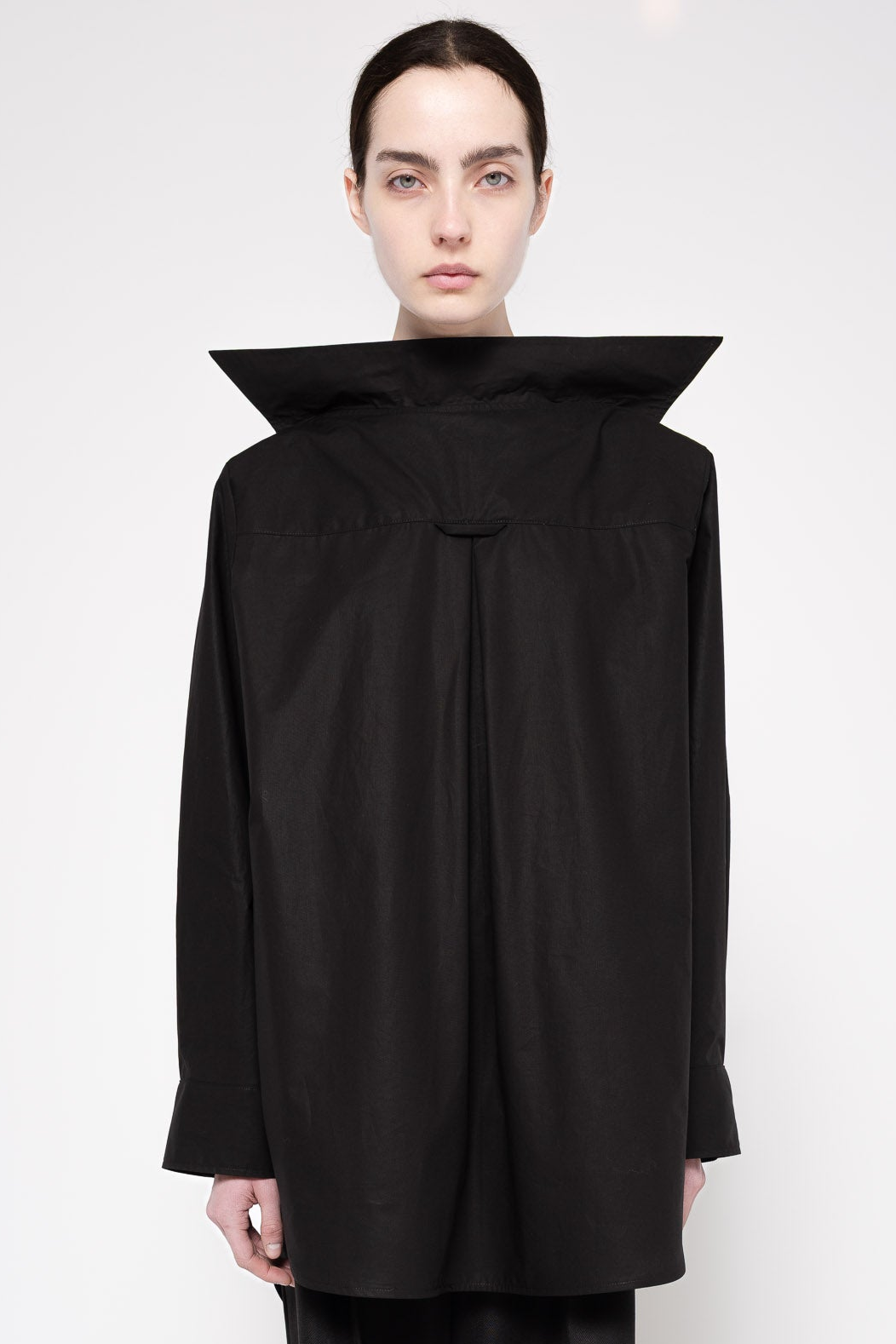A model wearing a black architectural long-sleeve top with a dramatic high collar and gathered front, photographed against a plain light gray studio background.