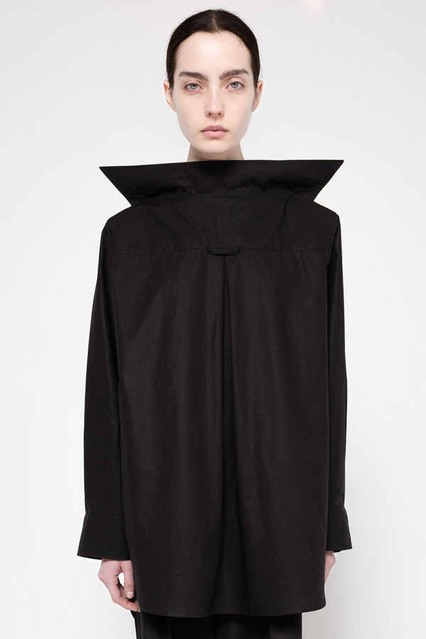 A model wearing a black architectural long-sleeve top with a dramatic high collar and gathered front, photographed against a plain light gray studio background.