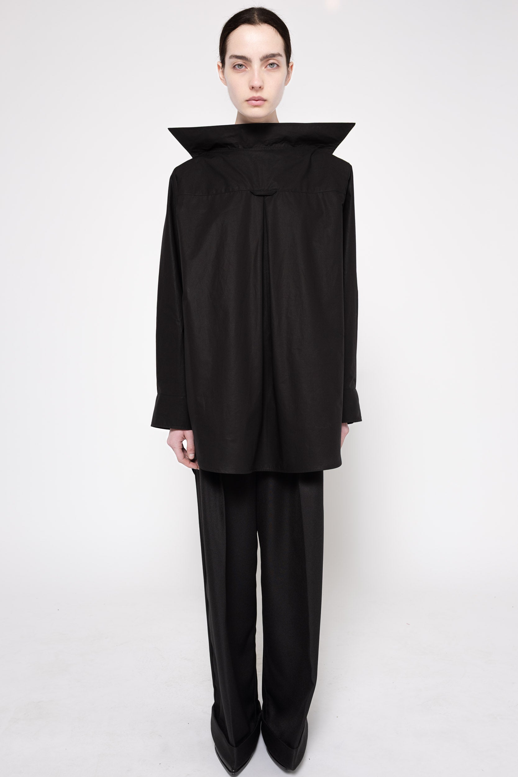 A model wearing a black asymmetrical long-sleeve top with a dramatic high collar and matching black wide-leg trousers, photographed against a seamless white studio background.