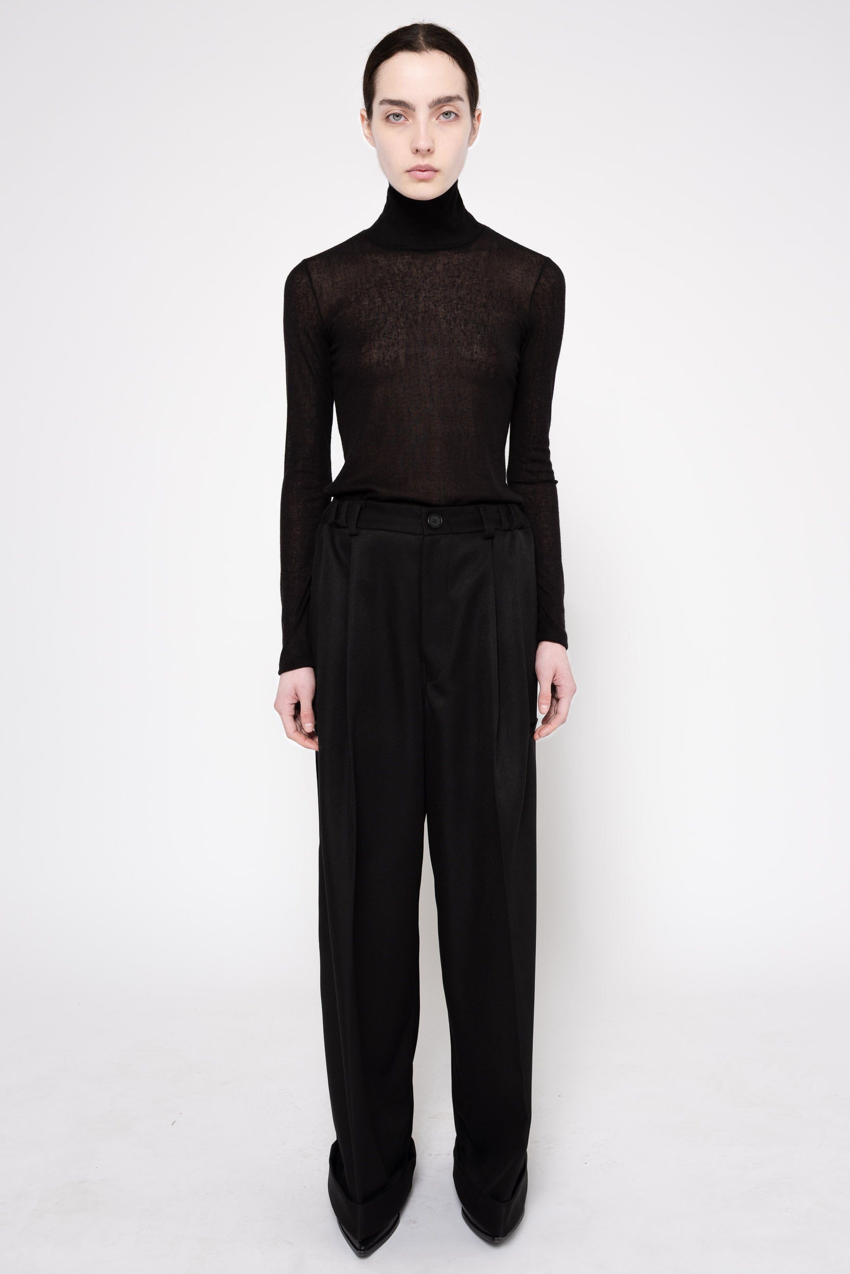 A model stands against a light gray studio background wearing a sheer black long-sleeve turtleneck top and high-waisted black wide-leg trousers.