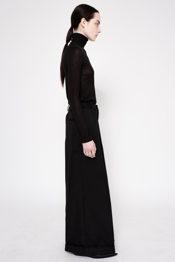 A model in profile wearing a black long-sleeve mock neck top and matching wide-leg trousers, photographed against a seamless white studio background.