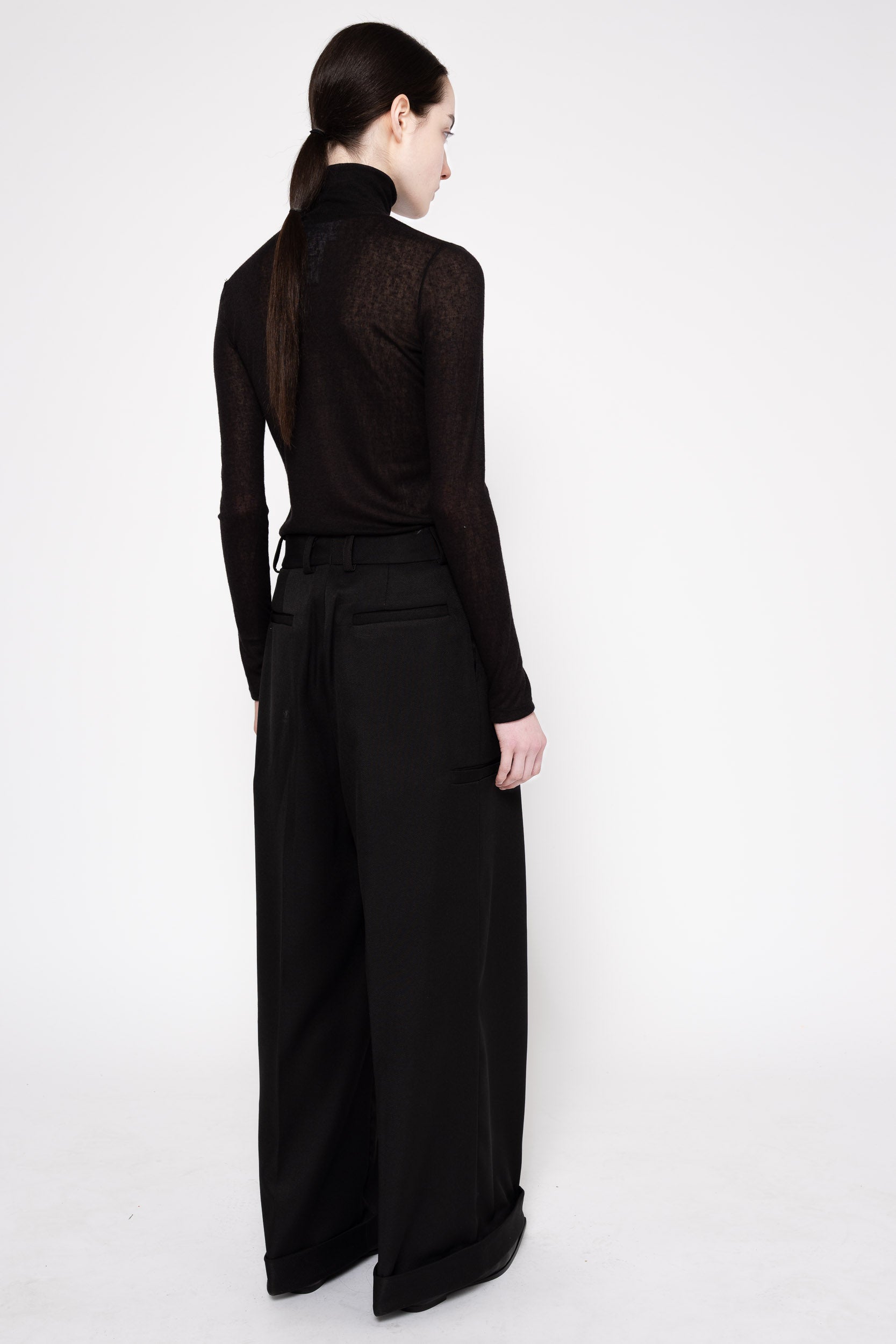 A model shown from the back wearing a black long-sleeve mock neck top with a sheer textured back panel and wide-leg black trousers, set against a plain white studio background.