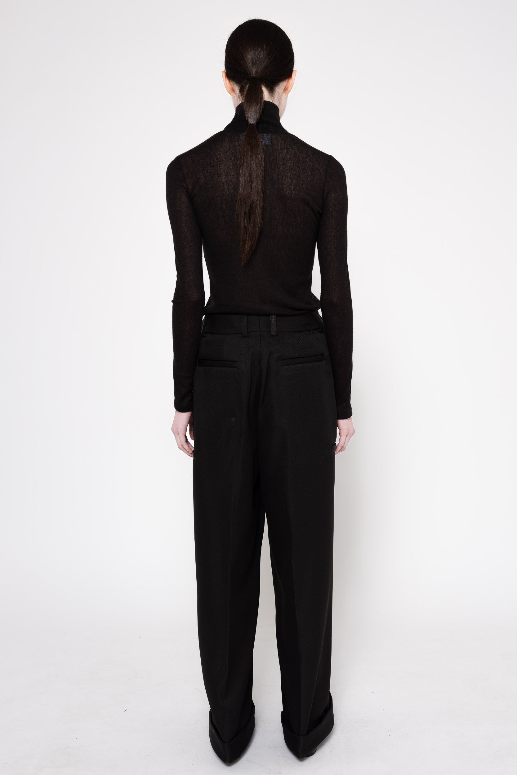 A model shown from the back wearing a black long-sleeve mock-neck knit top with sheer lace detailing and black wide-leg trousers, set against a plain white studio background.