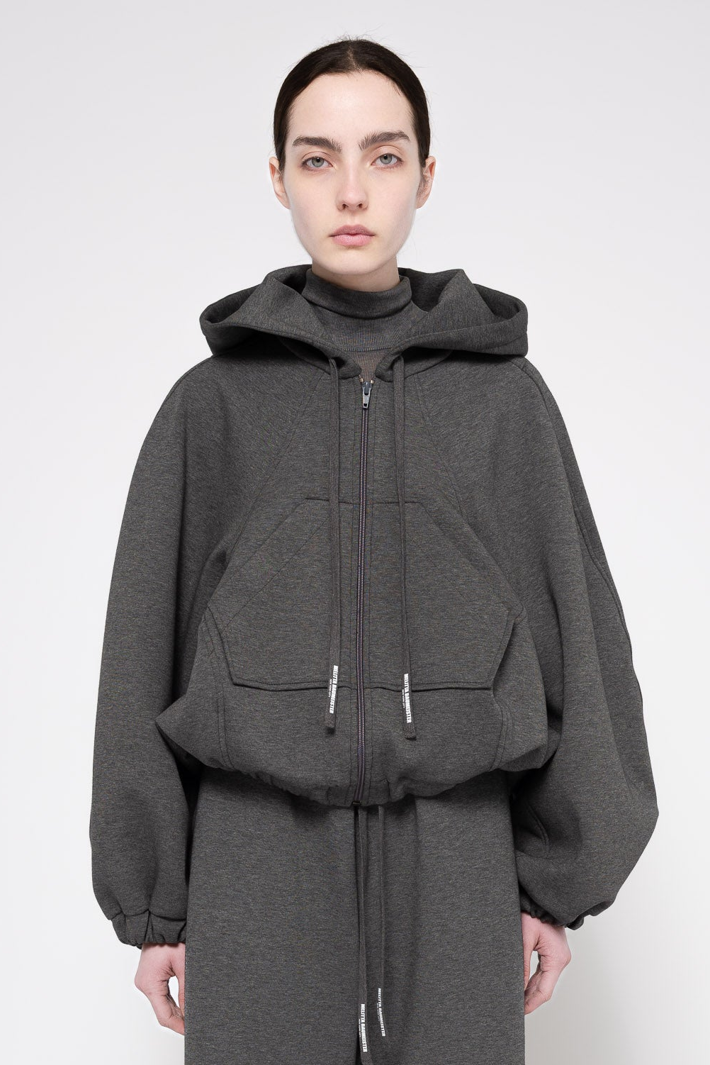 A model wearing a dark gray oversized zip-up hoodie with a hood, large front patch pockets, and voluminous balloon sleeves, paired with a matching dark gray drawstring skirt against a light gray studio background.