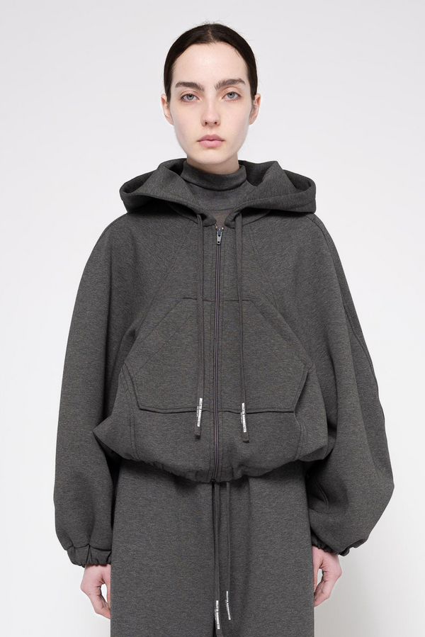 A model wearing a dark gray oversized zip-up hoodie with a hood, large front patch pockets, and voluminous balloon sleeves, paired with a matching dark gray drawstring skirt against a light gray studio background.