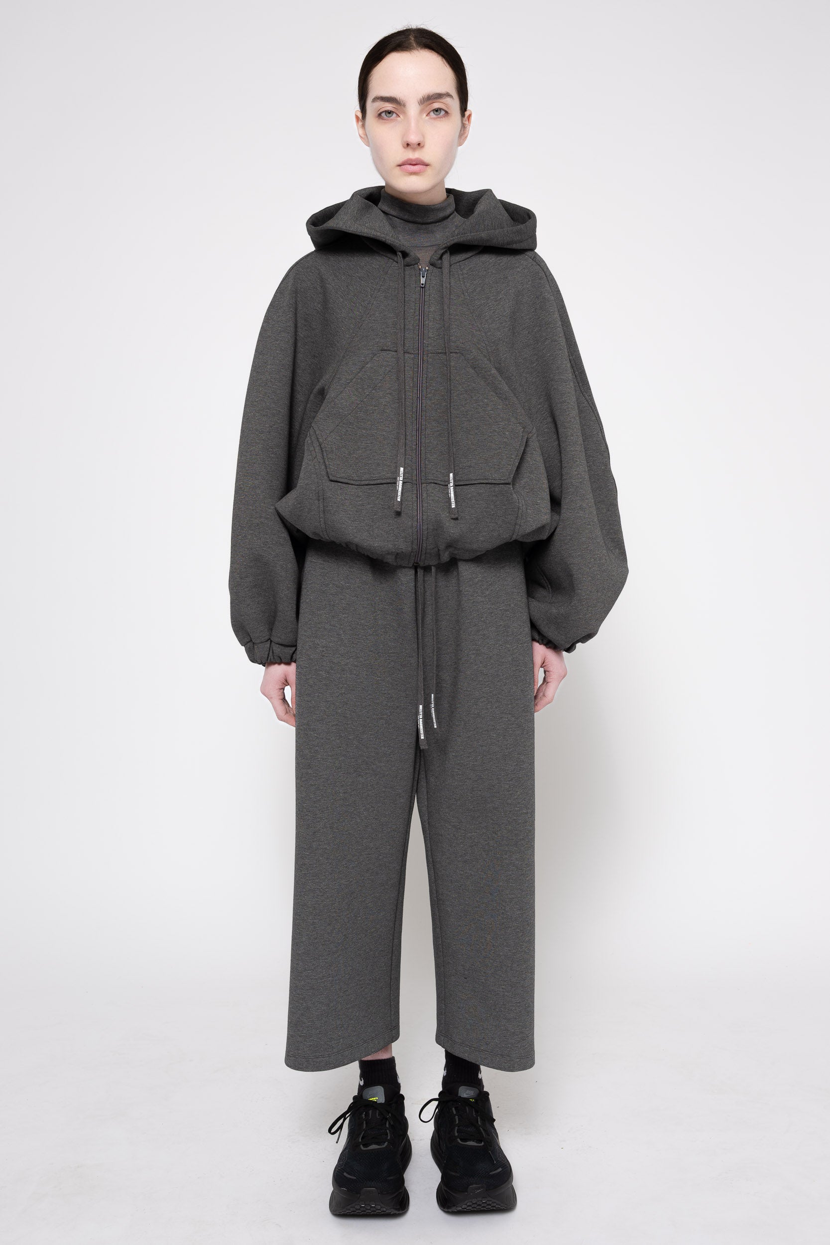 A model stands against a light gray studio background wearing a dark gray hooded zip-up jacket with oversized sleeves and matching wide-leg cropped pants, paired with black sneakers.