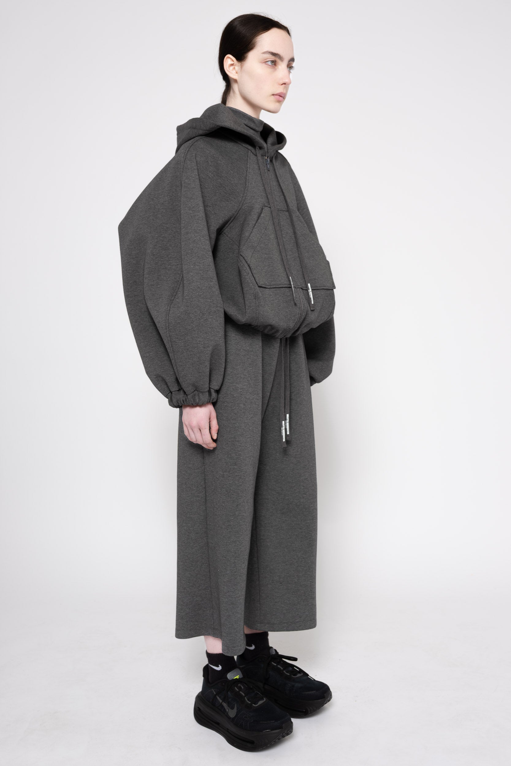 A model wearing a dark gray oversized hooded jacket with voluminous balloon sleeves and a matching long wide-leg skirt, styled with black sneakers against a plain light gray studio background.