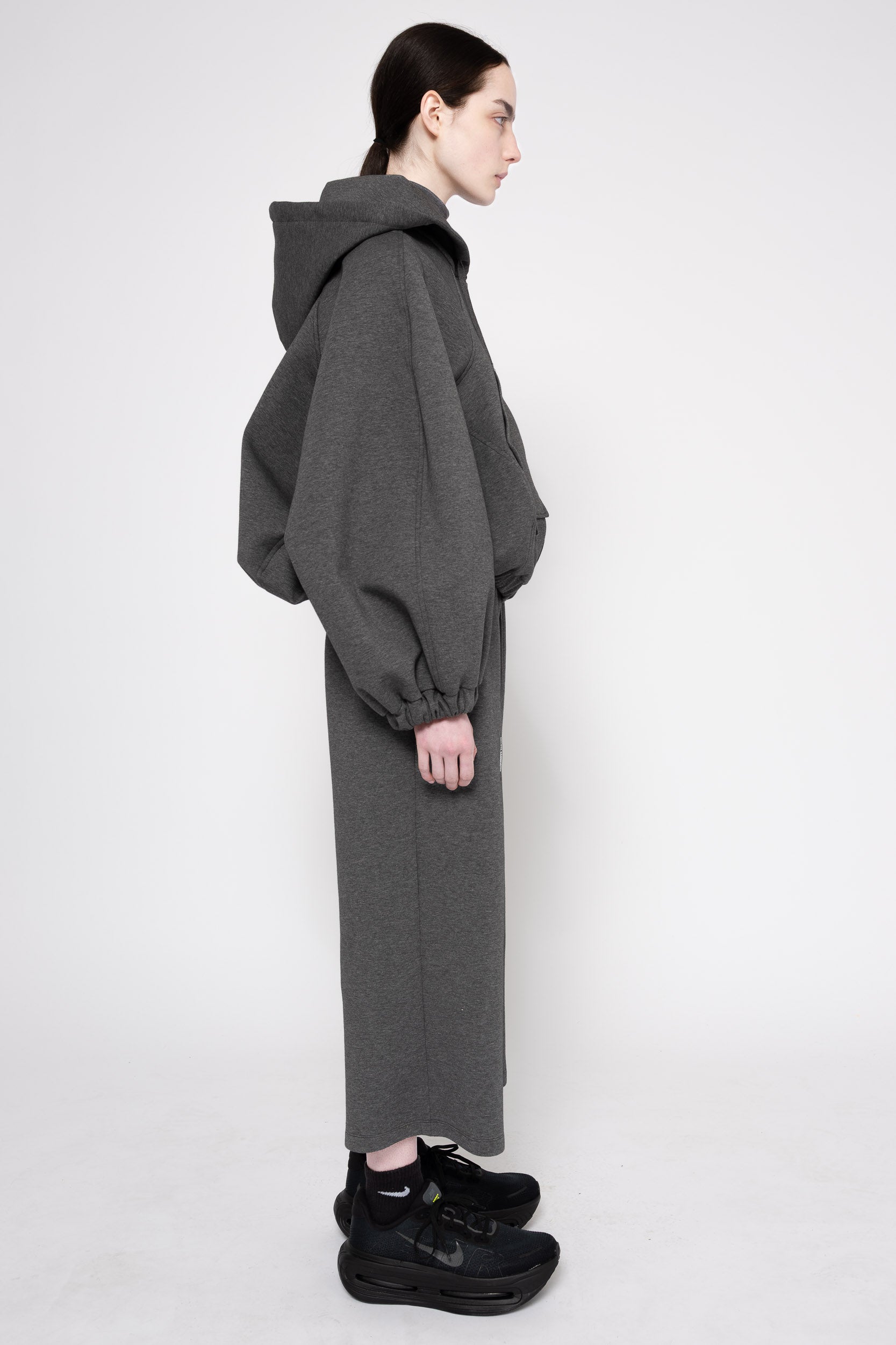 A model shown in profile wearing a dark gray oversized hooded sweatshirt and matching long skirt, paired with black sneakers, against a plain light gray studio background.