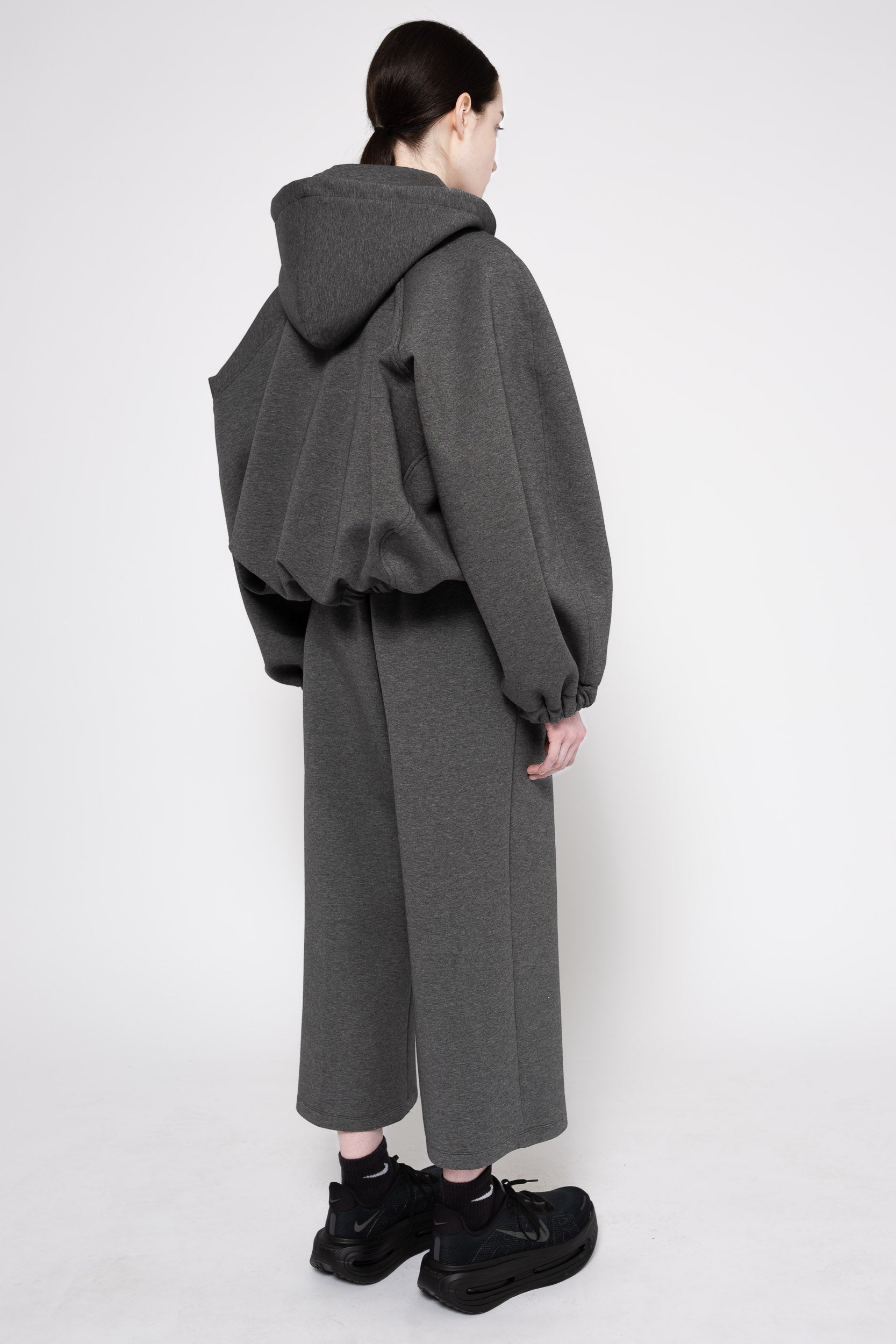 A model shown from the back wearing a dark gray oversized hooded sweatshirt with voluminous draped sleeves and matching wide-leg cropped trousers, paired with black boots against a plain light gray studio background.