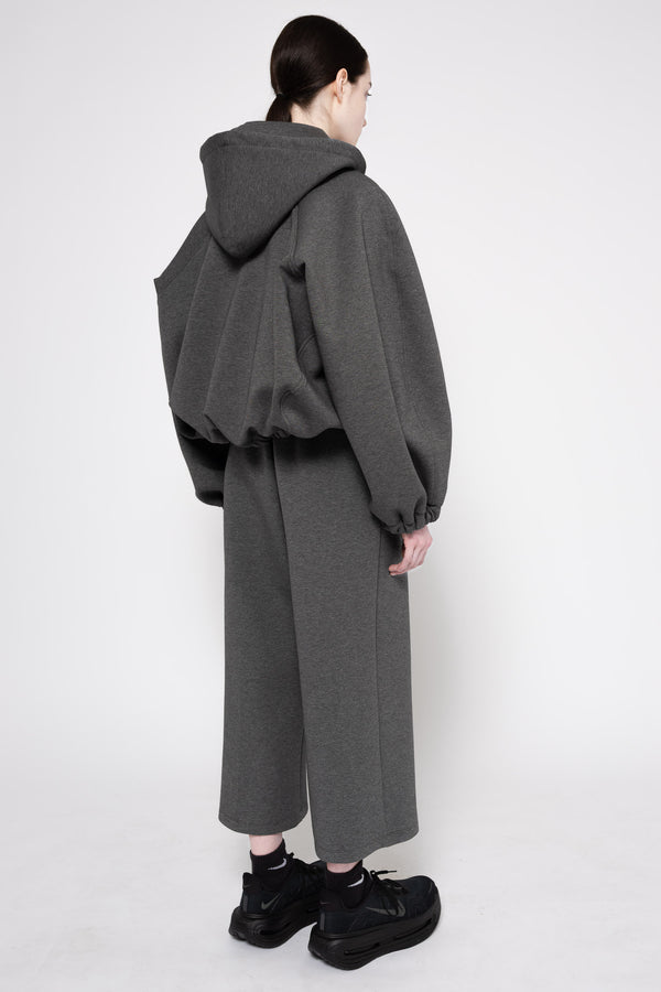 A model shown from the back wearing a dark gray oversized hooded sweatshirt with voluminous draped sleeves and matching wide-leg cropped trousers, paired with black boots against a plain light gray studio background.