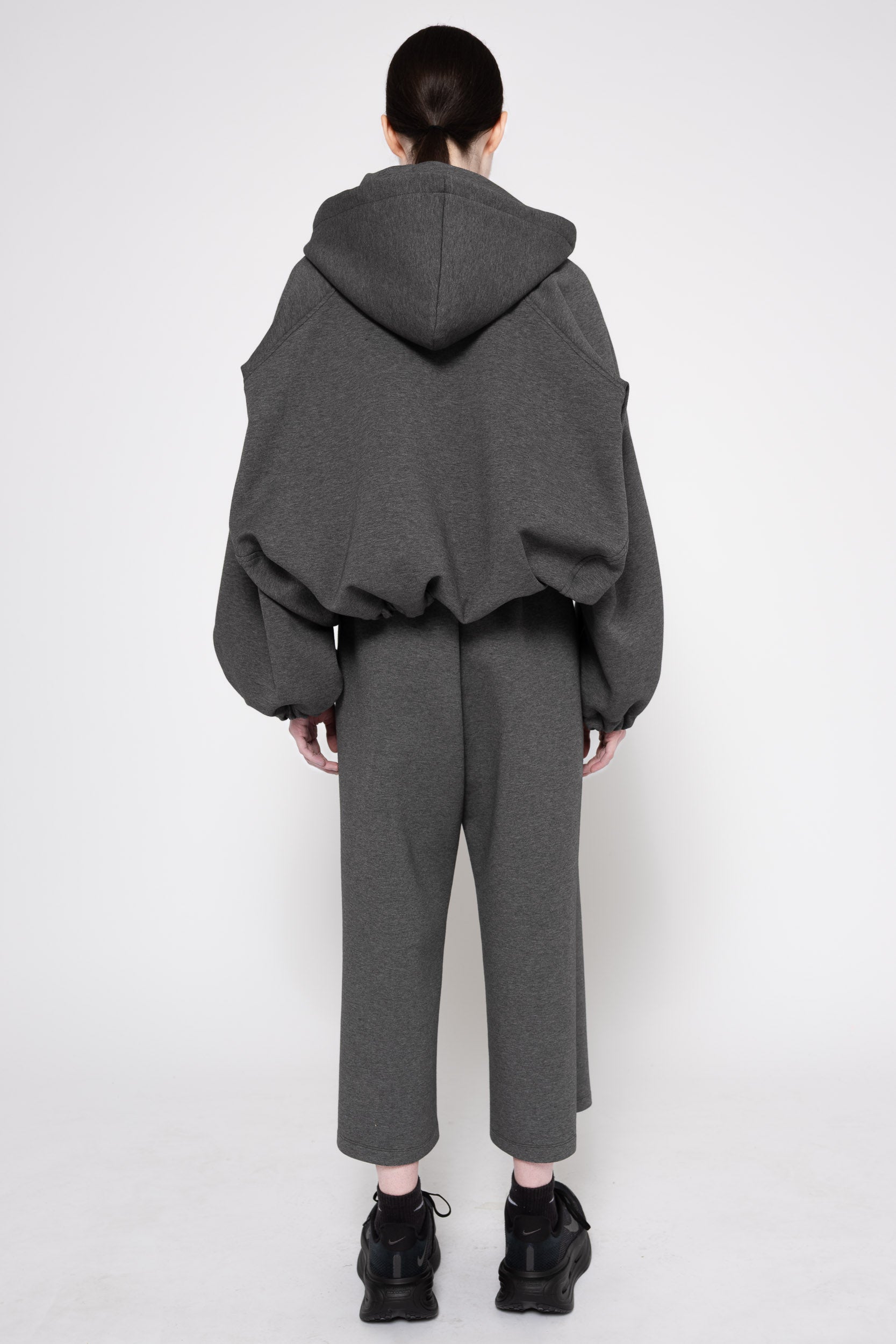 A model shown from the back wearing a dark gray oversized cropped hoodie and matching cropped trousers, paired with black sneakers, against a plain light gray studio background.