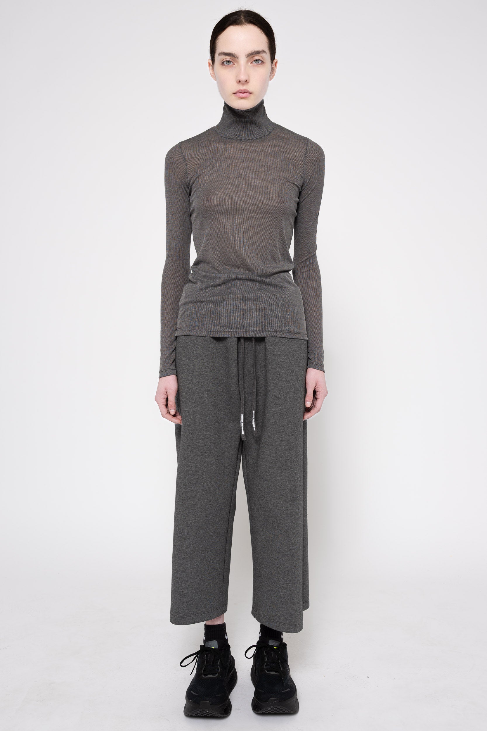 A model stands against a light gray studio background wearing a dark gray ribbed turtleneck sweater, matching wide-leg knit pants with a drawstring waist, black socks, and black sneakers.