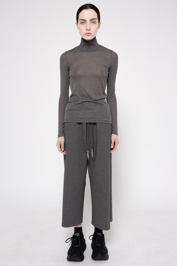 A model stands against a light gray studio background wearing a dark gray ribbed turtleneck sweater, matching wide-leg knit pants with a drawstring waist, black socks, and black sneakers.