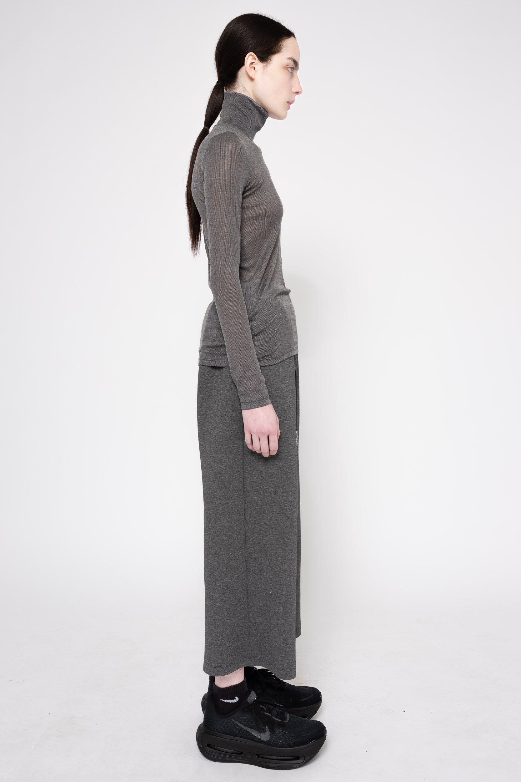 A model shown from the side wearing a dark gray ribbed turtleneck sweater and matching wide-leg cropped pants, paired with black sneakers, against a plain white studio background.