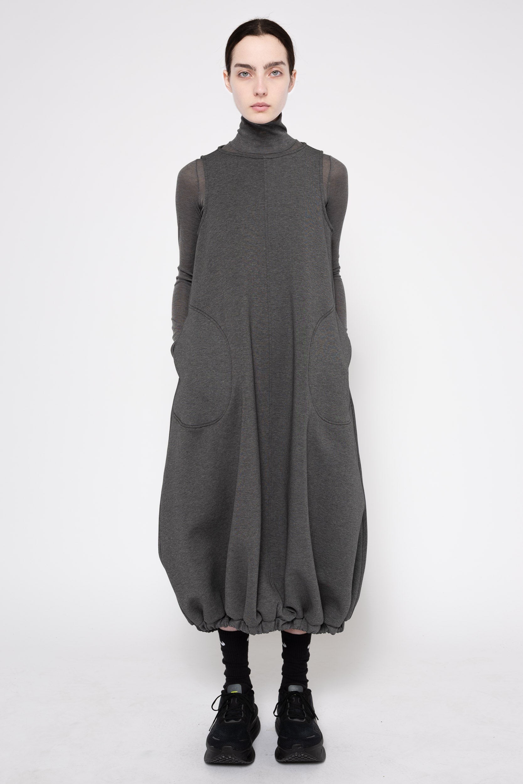 A model wearing a dark gray oversized turtleneck dress with sheer long sleeves and large front patch pockets, paired with black socks and black sneakers, against a plain light gray studio background.