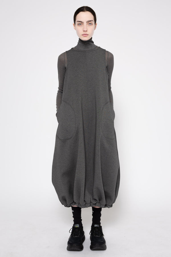 A model wearing a dark gray oversized turtleneck dress with sheer long sleeves and large front patch pockets, paired with black socks and black sneakers, against a plain light gray studio background.