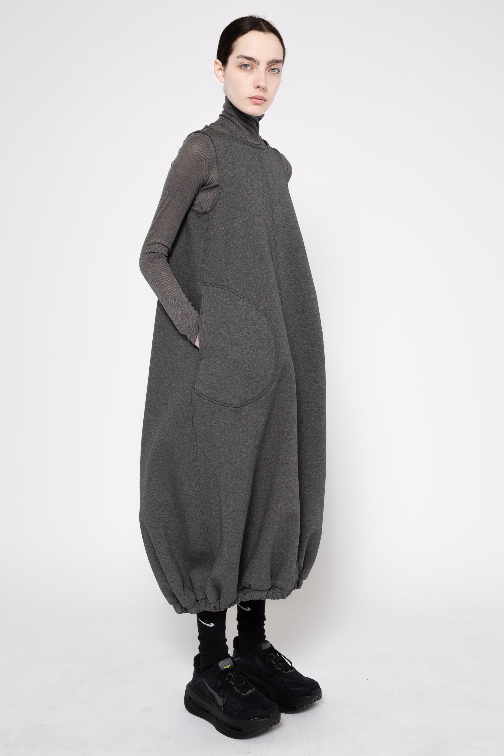 A model wearing a dark gray oversized long-sleeve turtleneck dress with voluminous balloon hems and large front patch pockets, styled with black socks and black sneakers against a plain light gray studio background.
