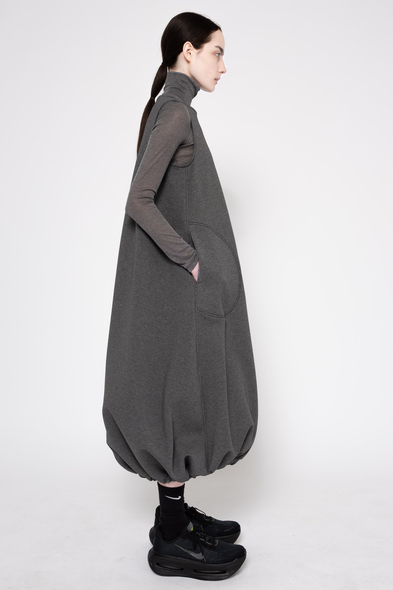 A model shown in profile wearing a dark gray oversized long-sleeve turtleneck dress with voluminous balloon hems and side pockets, styled with black socks and black sneakers against a plain light gray studio background.