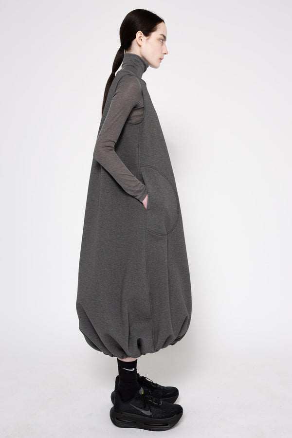 A model shown in profile wearing a dark gray oversized long-sleeve turtleneck dress with voluminous balloon hems and side pockets, styled with black socks and black sneakers against a plain light gray studio background.
