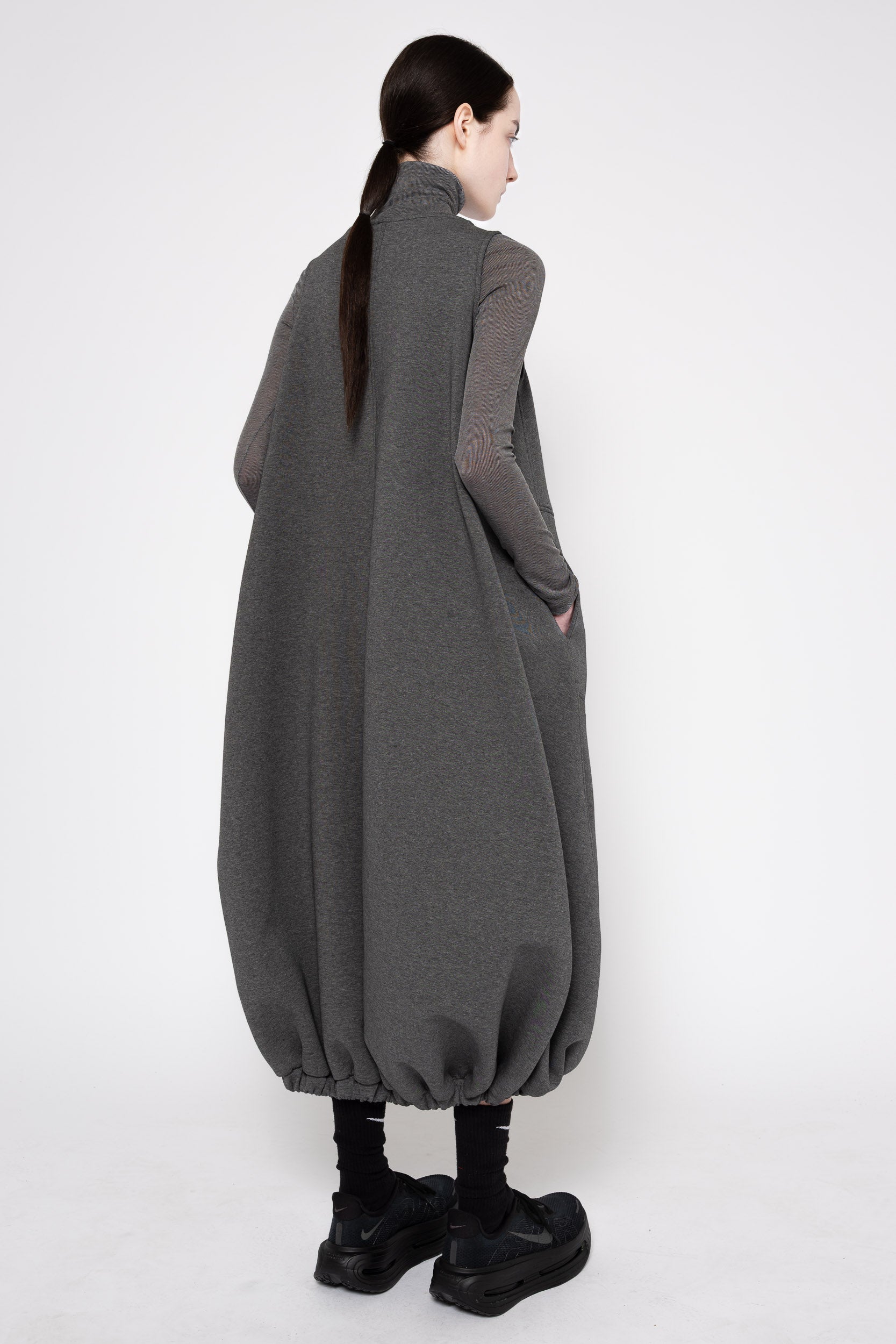 A model shown from the back wearing a dark gray oversized cape-style dress with long sheer sleeves and black platform shoes, photographed against a plain white studio background.