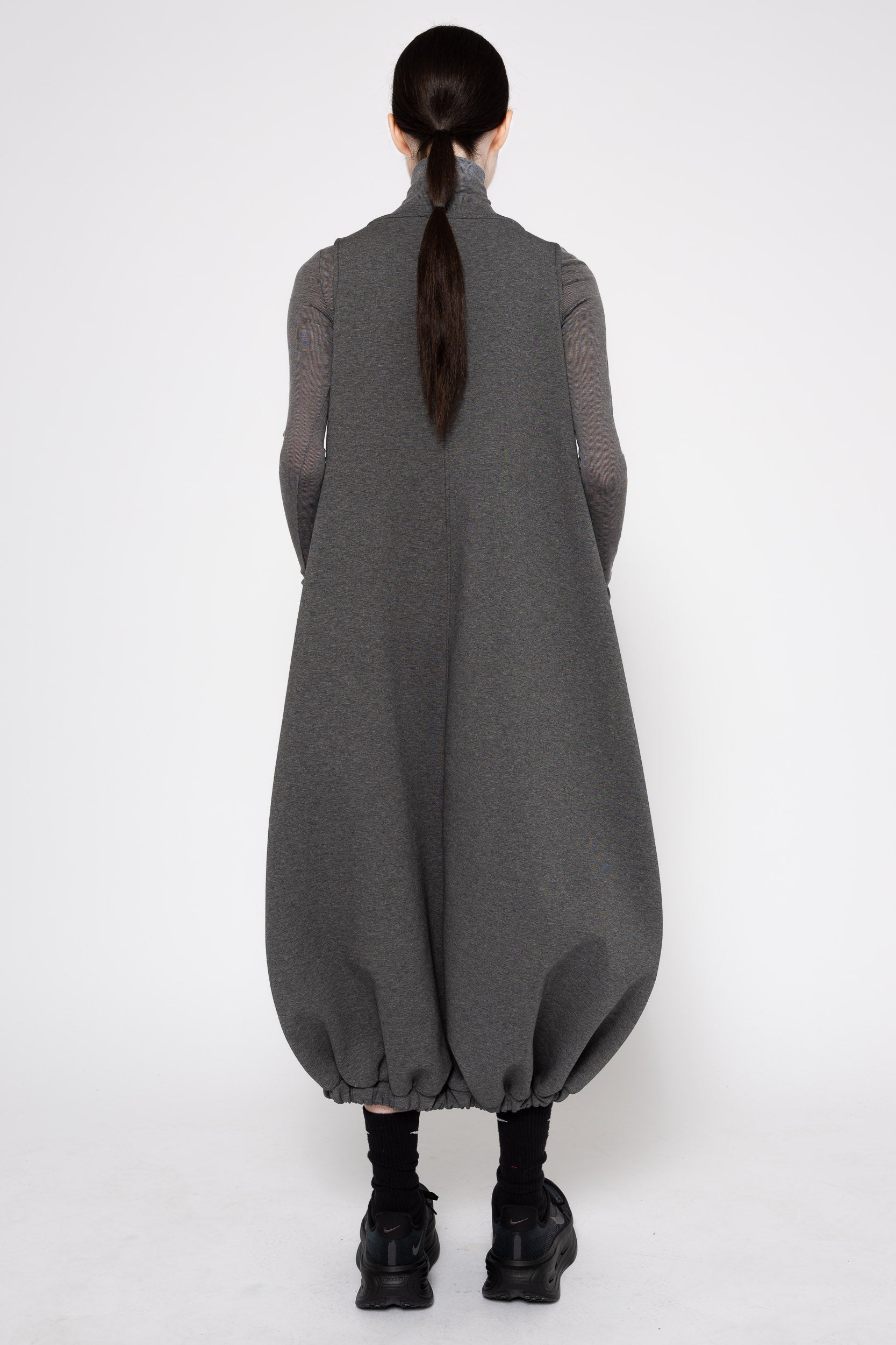 A model viewed from the back wearing a dark gray long-sleeve turtleneck dress with voluminous balloon hems and black boots, set against a plain light gray studio background.